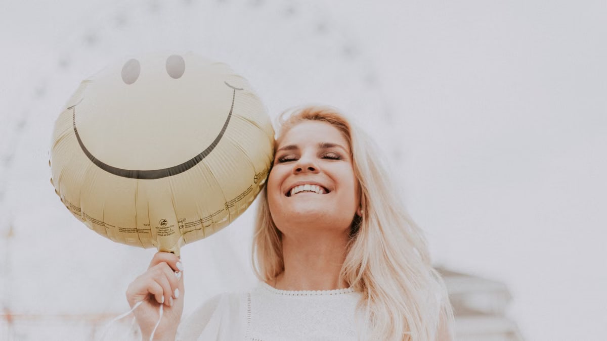 Cheerful woman holding a smiley balloon outdoors on a sunny day, exuding happiness and positivity.